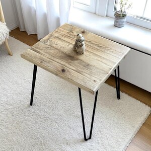 May include: Square wooden side table with a light brown, weathered finish. The tabletop is supported by black hairpin legs. A small decorative figurine sits on the table. The table is approximately 24 inches square.