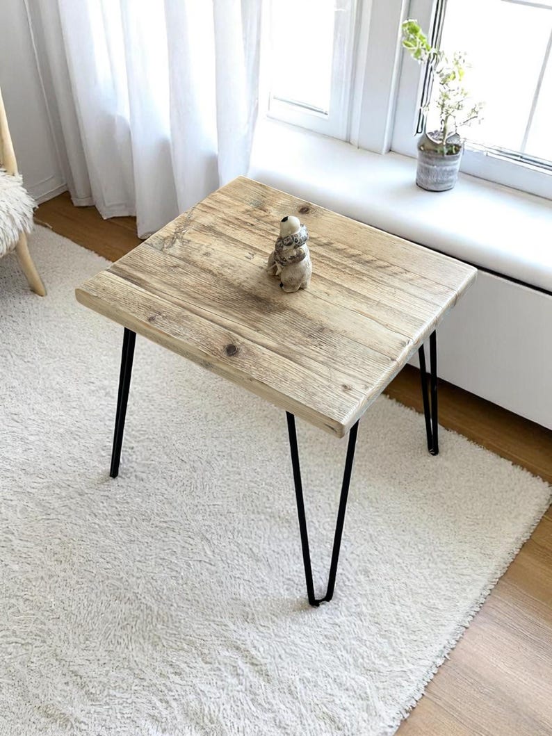 May include: Square wooden side table with a light brown, weathered finish. The tabletop is supported by black hairpin legs. A small decorative figurine sits on the table. The table is approximately 24 inches square.