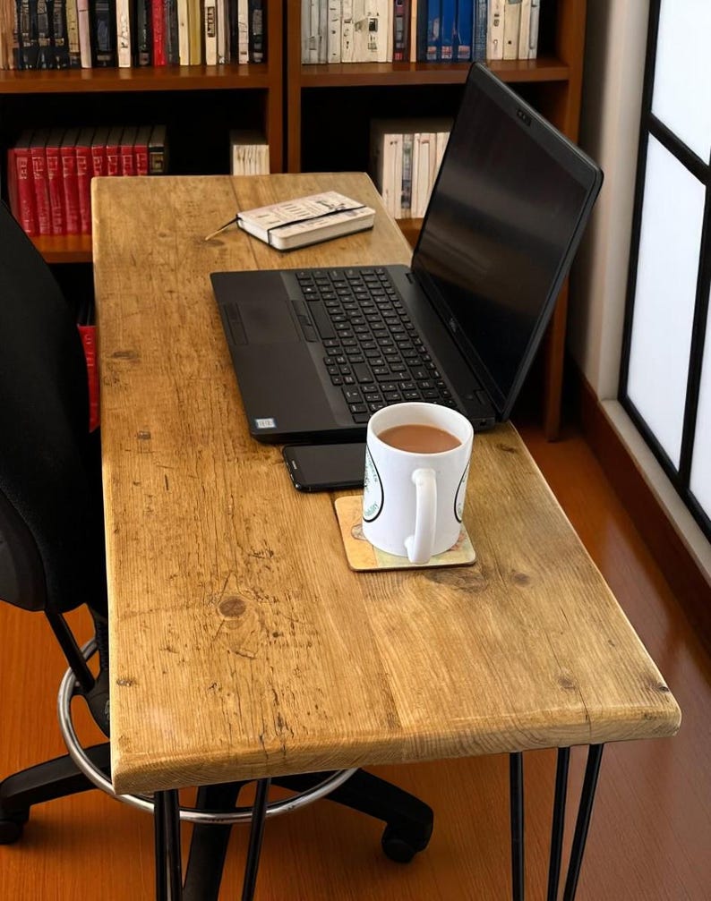 May include: A rectangular desk with a light brown wood grain surface and four black metal hairpin legs. The desk holds a black laptop, a small notebook, a black smartphone, and a white ceramic mug filled with a brown beverage on a coaster.