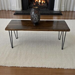 May include: A rectangular coffee table with a dark stained wooden top and black hairpin legs. A decorative metal vase with a leaf pattern sits on the table. The table is placed on a white rug in front of a fireplace.