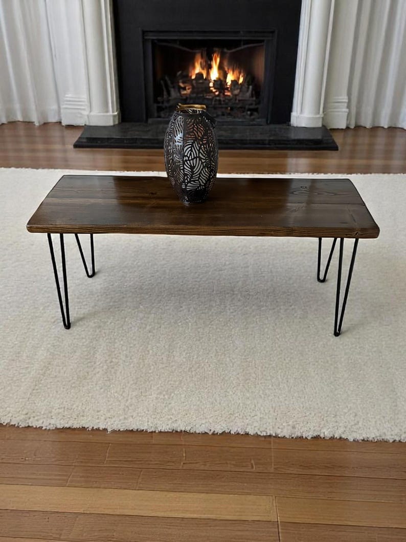 May include: A rectangular coffee table with a dark stained wooden top and black hairpin legs. A decorative metal vase with a leaf pattern sits on the table. The table is placed on a white rug in front of a fireplace.