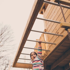 May include: A young girl in a pink and green striped jacket and blue jeans climbs a wooden ladder on a playground. She is looking up and has a surprised expression on her face.