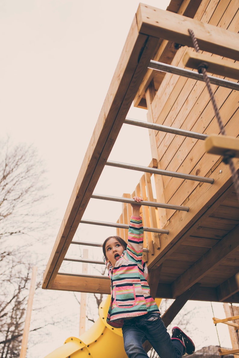 May include: A young girl in a pink and green striped jacket and blue jeans climbs a wooden ladder on a playground. She is looking up and has a surprised expression on her face.