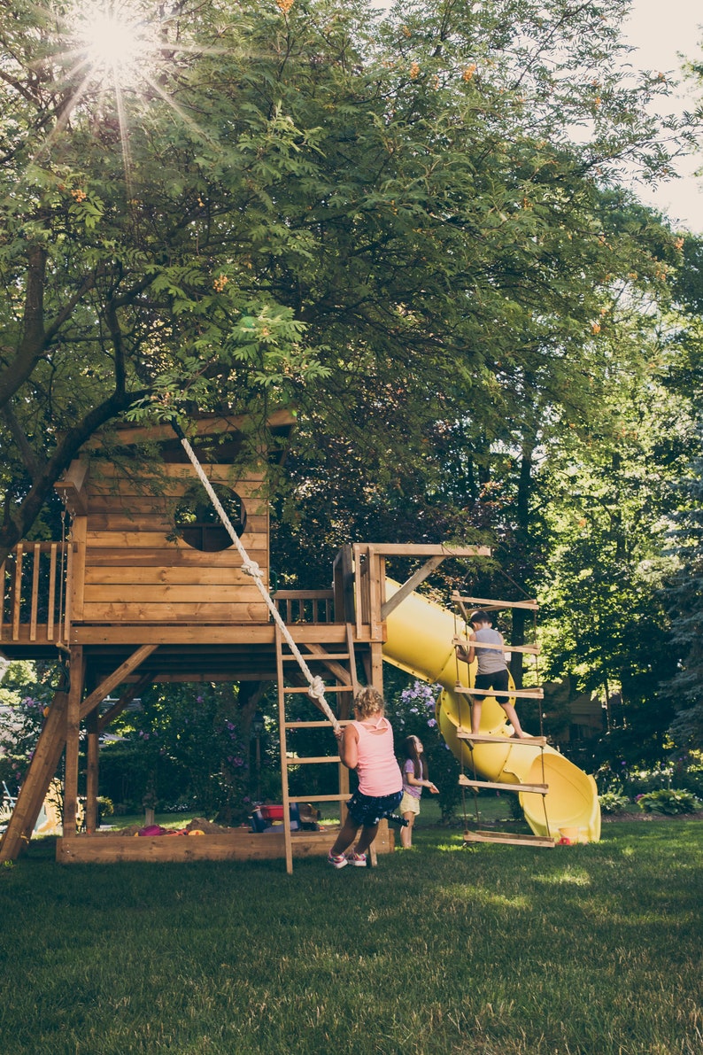 May include: A wooden treehouse with a yellow slide and a rope ladder. Two children are playing on the treehouse and slide.