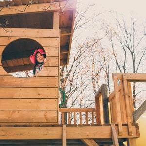 May include: A young girl wearing a pink hat peeks out of a circular window in a wooden playhouse. The playhouse has a wooden deck and railing. The background is a blurred image of trees.