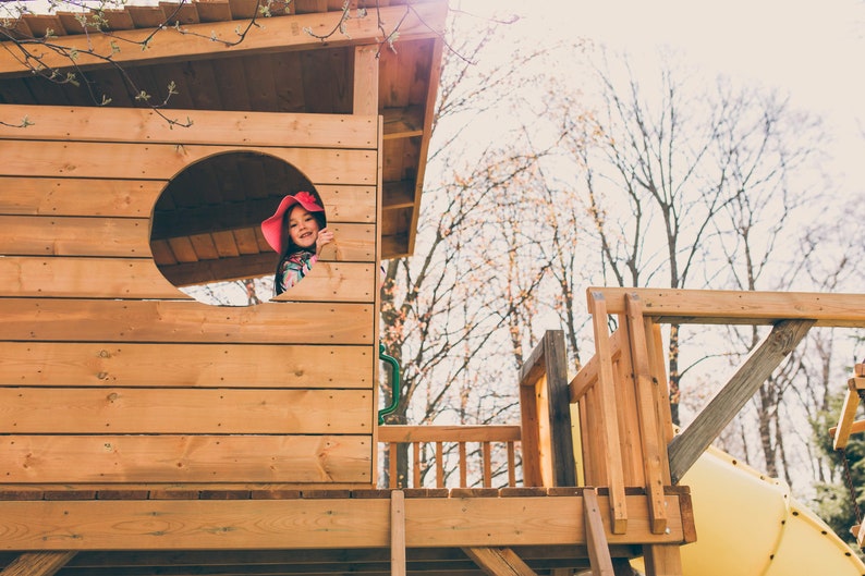 May include: A young girl wearing a pink hat peeks out of a circular window in a wooden playhouse. The playhouse has a wooden deck and railing. The background is a blurred image of trees.