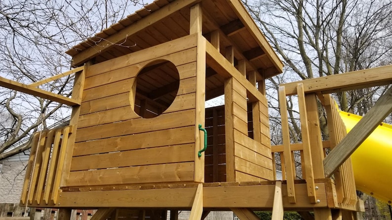 May include: A wooden playhouse with a roof, a window, and a door. The playhouse is made of natural wood and has a green door handle.