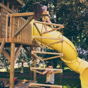 May include: Two children play on a wooden playground set with a yellow slide. One child climbs a rope ladder while the other sits on a wooden platform.