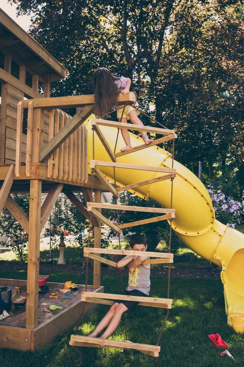 May include: Two children play on a wooden playground set with a yellow slide. One child climbs a rope ladder while the other sits on a wooden platform.