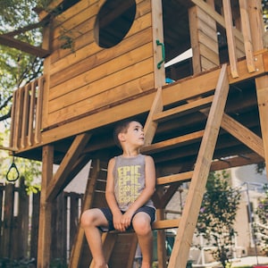 May include: A young boy wearing a grey tank top with the word "EPIC" printed on it sits on a wooden ladder attached to a wooden playhouse. The playhouse has a large window with a circular opening.