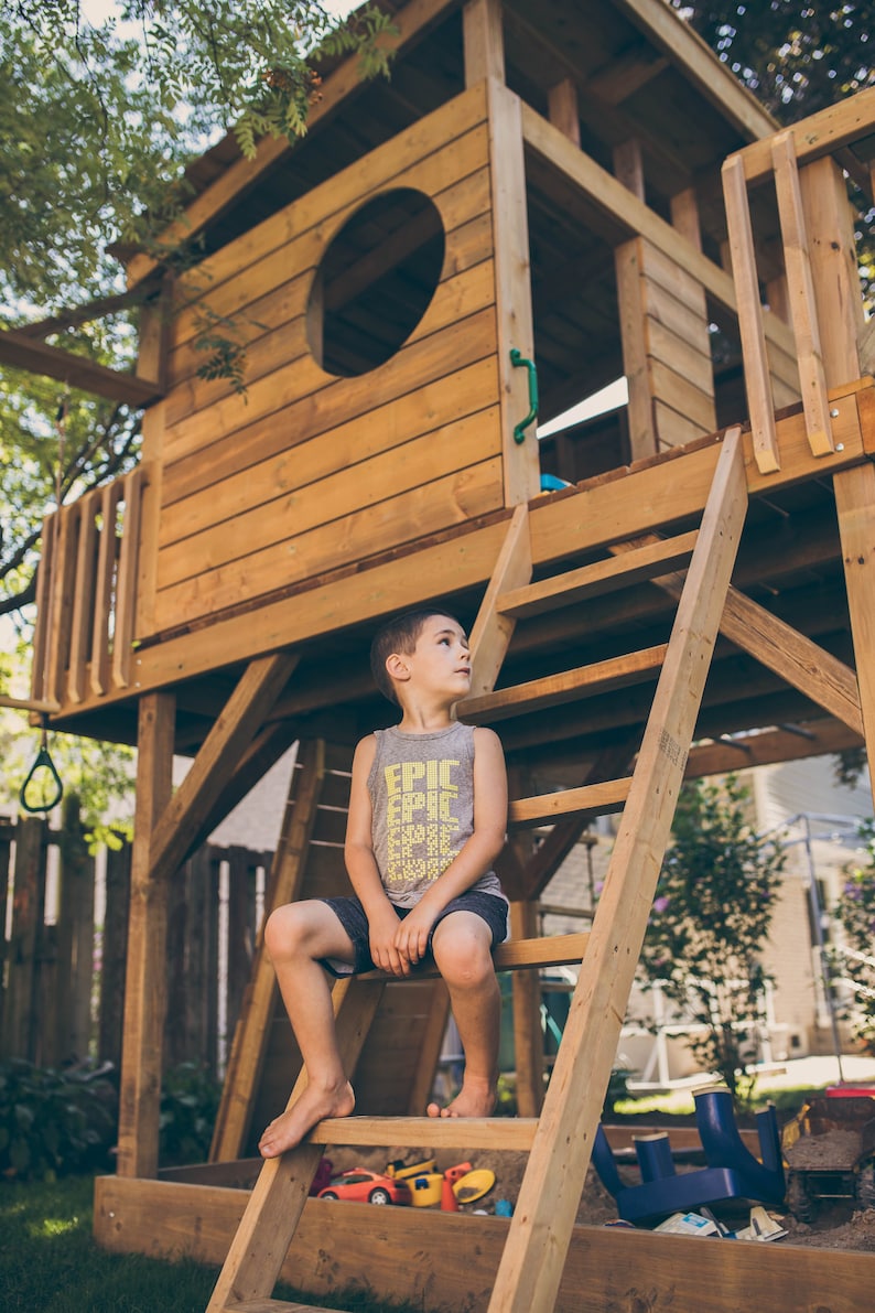 May include: A young boy wearing a grey tank top with the word "EPIC" printed on it sits on a wooden ladder attached to a wooden playhouse. The playhouse has a large window with a circular opening.