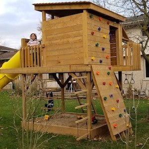 May include: A wooden playset with a slide, a climbing wall, and a platform. The playset is painted brown and has a yellow slide. A child is standing on the platform.