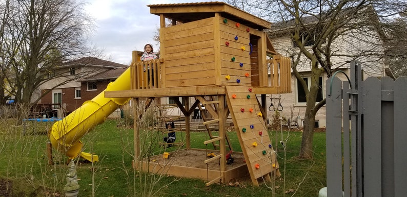 May include: A wooden playset with a slide, a climbing wall, and a platform. The playset is painted brown and has a yellow slide. A child is standing on the platform.