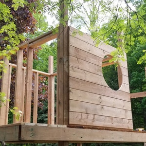 May include: A wooden treehouse under construction with a circular window opening. The treehouse is made of unfinished wood planks and beams. The treehouse is surrounded by green trees.