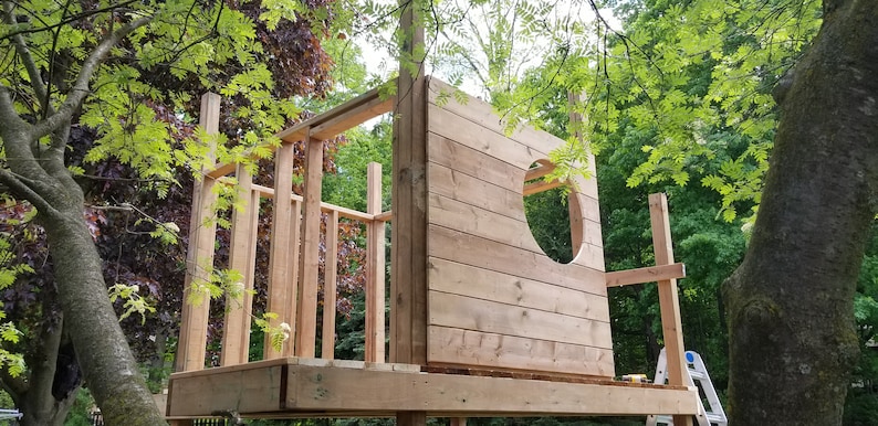 May include: A wooden treehouse under construction with a circular window opening. The treehouse is made of unfinished wood planks and beams. The treehouse is surrounded by green trees.