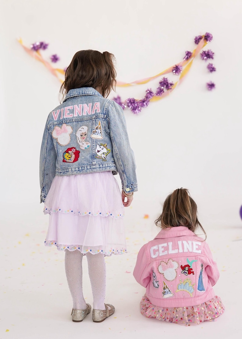 May include: Two children wearing denim and pink jackets with patches. The denim jacket has the name "VIENNA" on the back and Disney-themed patches. The pink jacket has the name "CELINE" on the back and Disney-themed patches. Both children are wearing skirts.