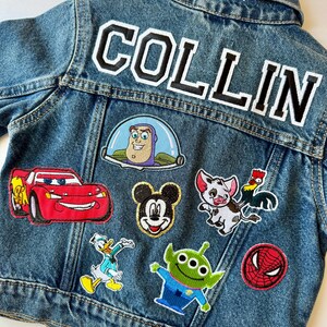 May include: A blue denim jacket featuring the name "COLLIN" in large, white-outlined black letters on the back. The jacket is adorned with various character patches, such as Mickey Mouse and Buzz Lightyear.