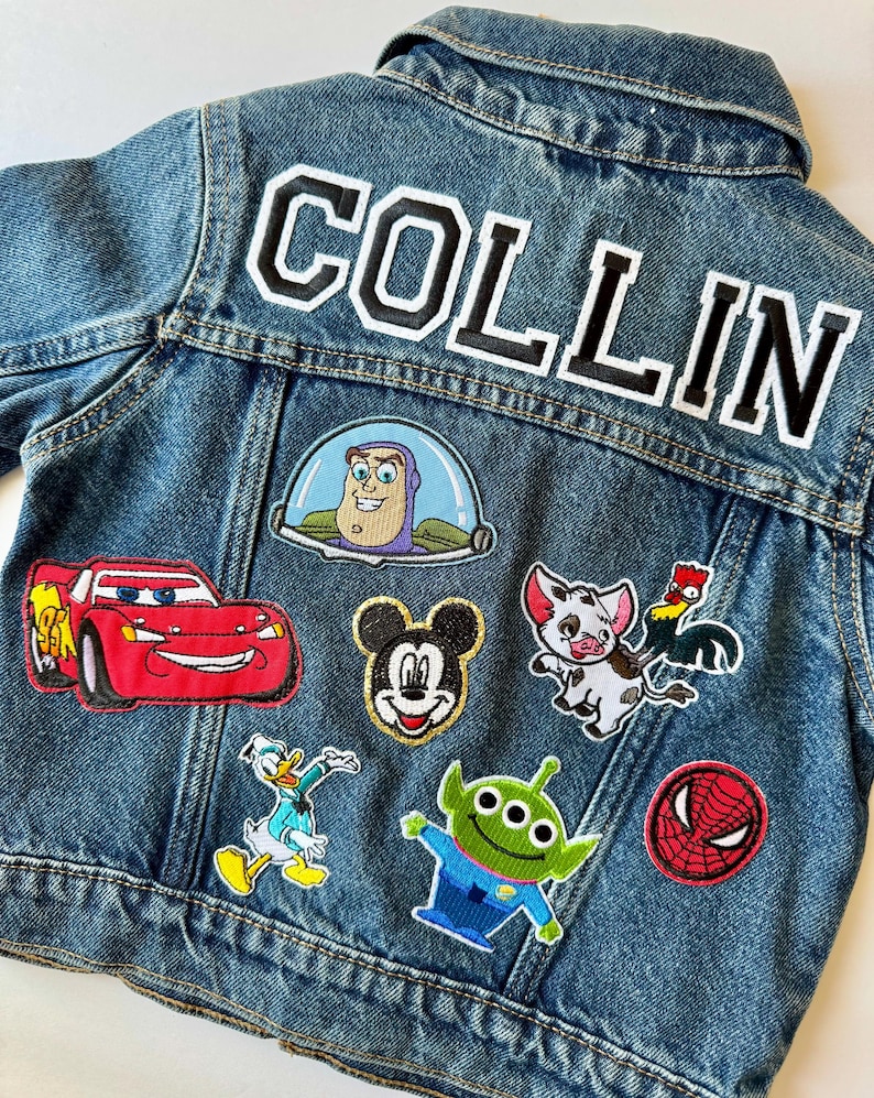 May include: A blue denim jacket featuring the name "COLLIN" in large, white-outlined black letters on the back. The jacket is adorned with various character patches, such as Mickey Mouse and Buzz Lightyear.