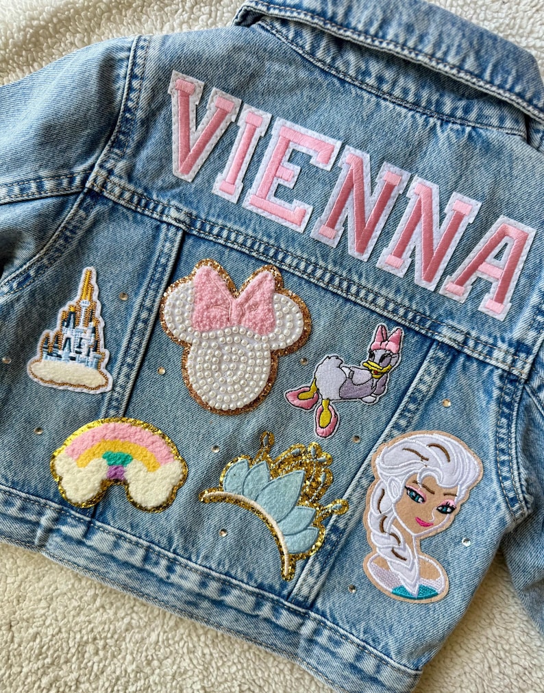 Personalized light-wash denim jacket featuring princess-themed patches, a rainbow, gold crown, and pink varsity-style name embroidery. Finished with sparkling rhinestones. Perfect for toddlers and kids as a birthday gift or magical outfit for a theme park princess trip.