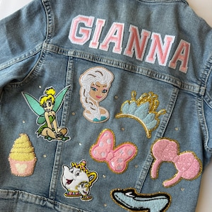Personalized light-wash denim jacket featuring princess-themed patches, a rainbow, gold crown, and pink varsity-style name embroidery. Finished with sparkling rhinestones. Perfect for toddlers and kids as a birthday gift or magical outfit for a theme park princess trip.