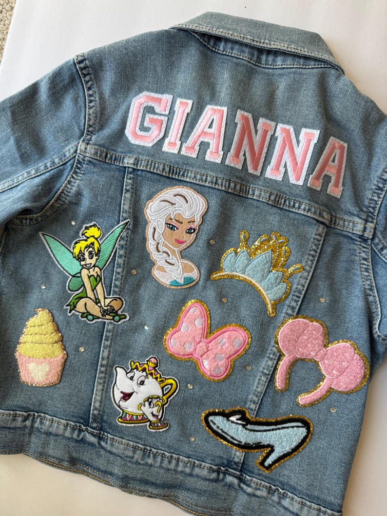 Personalized light-wash denim jacket featuring princess-themed patches, a rainbow, gold crown, and pink varsity-style name embroidery. Finished with sparkling rhinestones. Perfect for toddlers and kids as a birthday gift or magical outfit for a theme park princess trip.