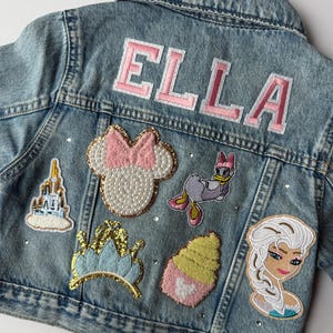 Personalized light-wash denim jacket featuring princess-themed patches, a rainbow, gold crown, and pink varsity-style name embroidery. Finished with sparkling rhinestones. Perfect for toddlers and kids as a birthday gift or magical outfit for a theme park princess trip.