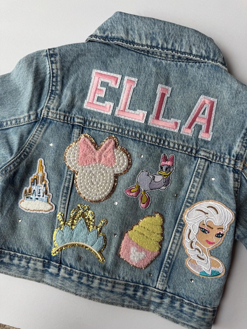 Personalized light-wash denim jacket featuring princess-themed patches, a rainbow, gold crown, and pink varsity-style name embroidery. Finished with sparkling rhinestones. Perfect for toddlers and kids as a birthday gift or magical outfit for a theme park princess trip.