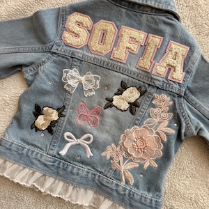 May include: Light blue denim jacket with the name "SOFIA" in large, cream-colored, varsity-style letters on the back. The jacket is decorated with embroidered flowers, butterflies, bows, and a ruffled hem.