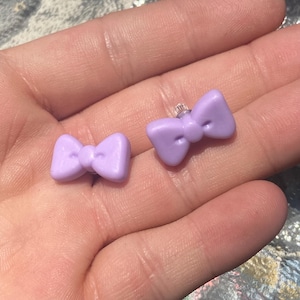 May include: A pair of light purple bow-shaped stud earrings. The earrings are made of a smooth material and are displayed on a hand. The bows have a classic, symmetrical design, with a small knot in the center.