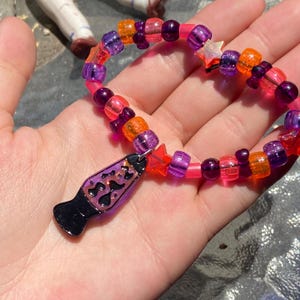 May include: A colorful beaded bracelet with a lava lamp charm. The bracelet features a mix of transparent and opaque beads in shades of pink, purple, and orange. The lava lamp charm is black with a purple and pink design, and the bracelet also includes star-shaped beads.