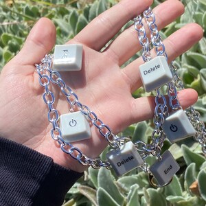 May include: A silver chain necklace featuring gray keyboard key pendants. The pendants include keys labeled "Delete", "End", and a power symbol. The necklace is held against a background of green foliage.