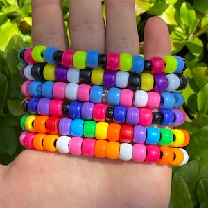 May include: A stack of colorful beaded bracelets in various shades of pink, purple, blue, green, yellow, and orange. The bracelets are made of small plastic beads and are stacked on top of each other.