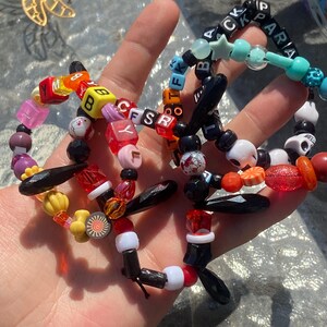 May include: A collection of colorful beaded bracelets. The bracelets feature a variety of beads, including lettered cubes, stars, skulls, and decorative shapes. The color palette includes red, orange, yellow, pink, black, and blue.