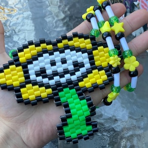 May include: A pixelated, yellow, black, white, and green beaded bracelet and cuff set. The cuff features a large, pixelated design, while the bracelet has yellow star and flower beads. The set is held in a hand.