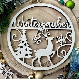 Può includere: Decorazione natalizia in legno con la scritta "Winterzauber". Il design include un albero di Natale, fiocchi di neve e una renna. La decorazione è posta su una fetta di legno con accenti festivi.