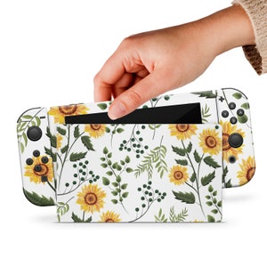 Può includere: Console Nintendo Switch bianca con una skin a motivi di girasoli e verde. La console è chiusa e lo schermo è visibile. La console è tenuta da una mano.
