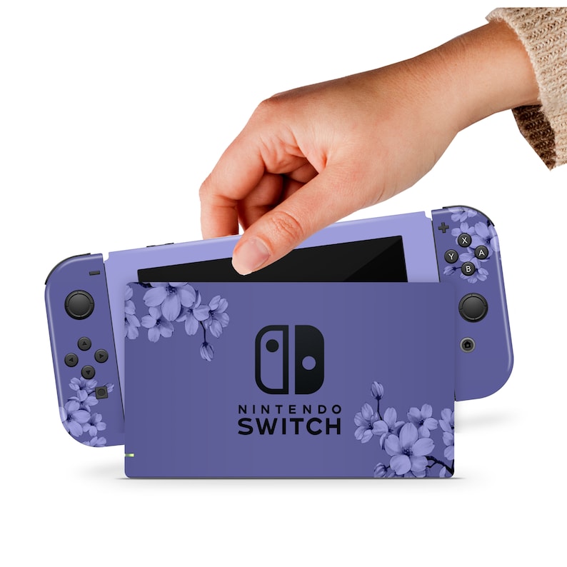 Puede incluir: Una consola Nintendo Switch morada con un dise&ntilde;o floral. La consola est&aacute; cerrada y la pantalla es negra. La consola est&aacute; sobre una superficie blanca.