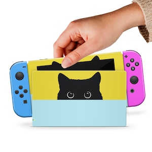 Può includere: Una console Nintendo Switch con controller Joy-Con blu e rosa. La console ha una skin gialla e azzurra con un gatto nero che sbuca da una striscia azzurra. La skin viene inserita nella console.