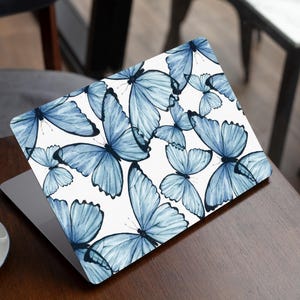 Könnte beinhalten: Ein Laptop mit weißem Hintergrund und einem Muster aus blauen Schmetterlingen. Die Schmetterlinge sind in einem zufälligen Muster angeordnet und haben alle unterschiedliche Größen.
