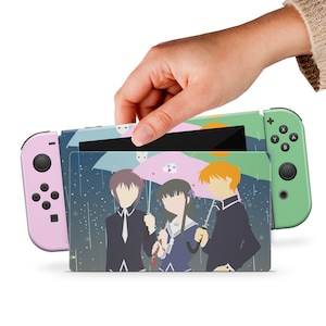 Pode incluir: Um console Nintendo Switch verde e rosa com uma capa personalizada que apresenta três personagens de anime segurando guarda-chuvas em um ambiente chuvoso. A capa tem um fundo azul com gotas de chuva brancas.