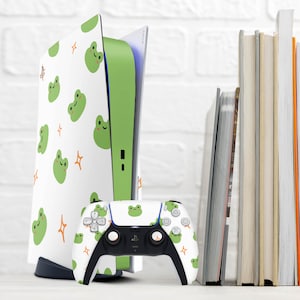 May include: A white and green video game console with a frog pattern and a matching white and green controller. The console is sitting on a white surface with a stack of books behind it.