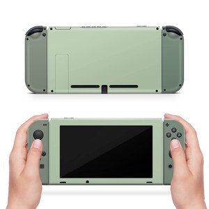 K&ouml;nnte beinhalten: Eine tragbare Videospielkonsole mit einem salbeigr&uuml;nen Mittelteil und abnehmbaren, dunkel olivgr&uuml;nen Controllern an beiden Seiten. Das Ger&auml;t verf&uuml;gt &uuml;ber einen schwarzen Bildschirm, Richtungstasten, Aktionstasten und ein r&uuml;ckseitiges L&uuml;ftungsgitter.
