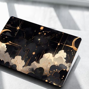 May include: A black laptop case featuring a celestial design with gold accents. The design includes crescent moons, stars, and cloud-like shapes. The case is on a light-colored surface next to a marble-patterned mug.