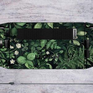May include: A Lenovo Legion handheld gaming device with a custom skin featuring a lush, dark green botanical design. The skin showcases various leaves, ferns, and white flowers, with the Lenovo logo visible.
