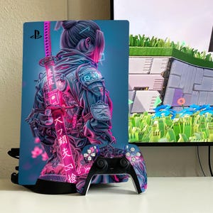 May include: A pink and blue video game console with a cyberpunk-style design featuring a woman with a katana sword. The console is on a white surface with a black controller.