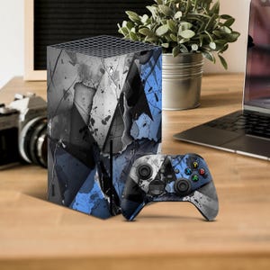 Peut inclure: Une console de jeu vidéo et une manette à motifs bleus, gris et noirs. La console a un design géométrique avec un aspect craquelé et vieilli. La manette a le même motif que la console.