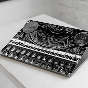 Könnte beinhalten: Ein schwarz-weißes Laptop mit einem Vintage-Schreibmaschinen-Design auf der Abdeckung. Die Tasten der Schreibmaschine sind sichtbar, darunter die Zahlen 1 bis 9, die Buchstaben QWERTYUIOP und die Symbole ~!@#$%^&*()_+=-`.