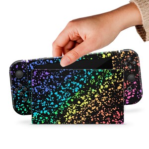 May include: A black Nintendo Switch console with a rainbow splatter paint design. The console is open and the screen is visible.