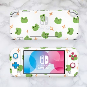May include: White Nintendo Switch Lite console with a custom skin featuring a repeating pattern of green frogs and orange stars. The console has a pink and blue screen and the text "NINTENDO SWITCH" is printed on the front.
