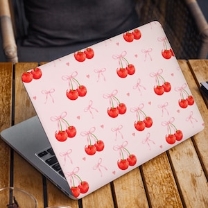 Può includere: Un laptop rosa con un motivo a ciliegie. Le ciliegie sono rosse e hanno fiocchi rosa. Il laptop è chiuso.